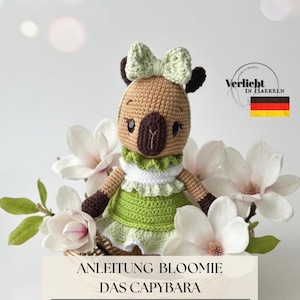 Könnte beinhalten: Eine gehäkelte Capybara-Puppe mit grünem Kleid und Schleife, in einem Korb mit weißen Blumen. Die Puppe ist aus braunem und beigem Garn mit einem weiß-grünen Rüschenkragen gefertigt. Der Text auf dem Bild lautet: "ANLEITUNG BLOOMIE DAS CAPYBARA".