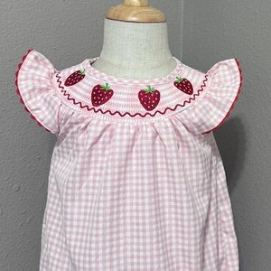 K&ouml;nnte beinhalten: Ein rosa-wei&szlig;es Gingham-Baby-Bodysuit mit gestickten Erdbeeren um den Halsausschnitt. Der Bodysuit hat kurze, gekr&auml;uselte &Auml;rmel. Der Bodysuit befindet sich auf einer Holzpuppe.