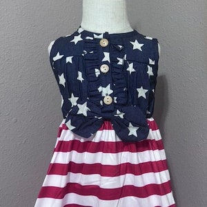 May include: A sleeveless dress with a blue and white star print top and a red and white striped skirt. The dress is on a mannequin and has a bow at the waist.