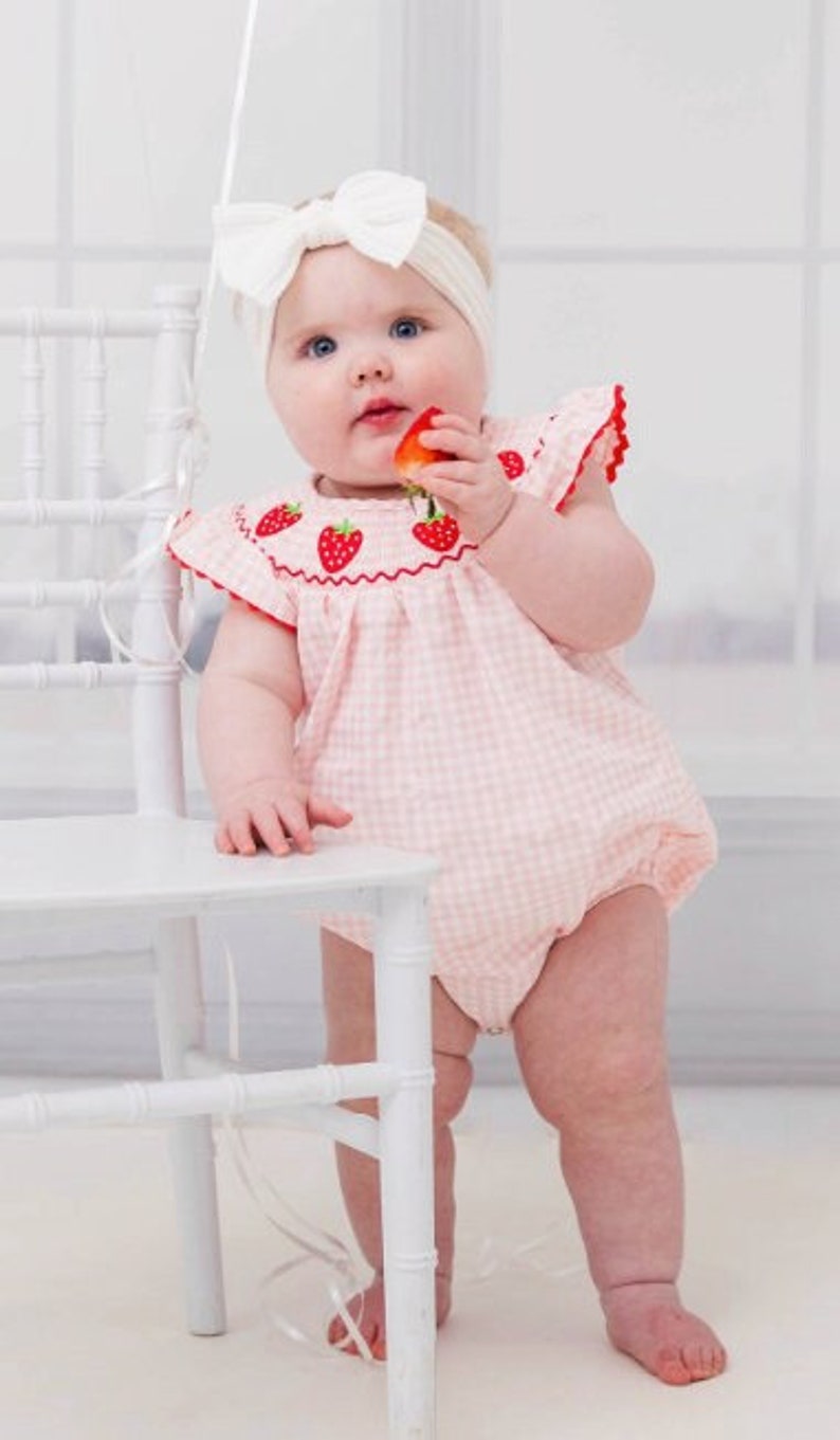May include: A baby girl wearing a pink and white gingham dress with embroidered strawberries. She is holding a strawberry in her hand and is standing next to a white chair.