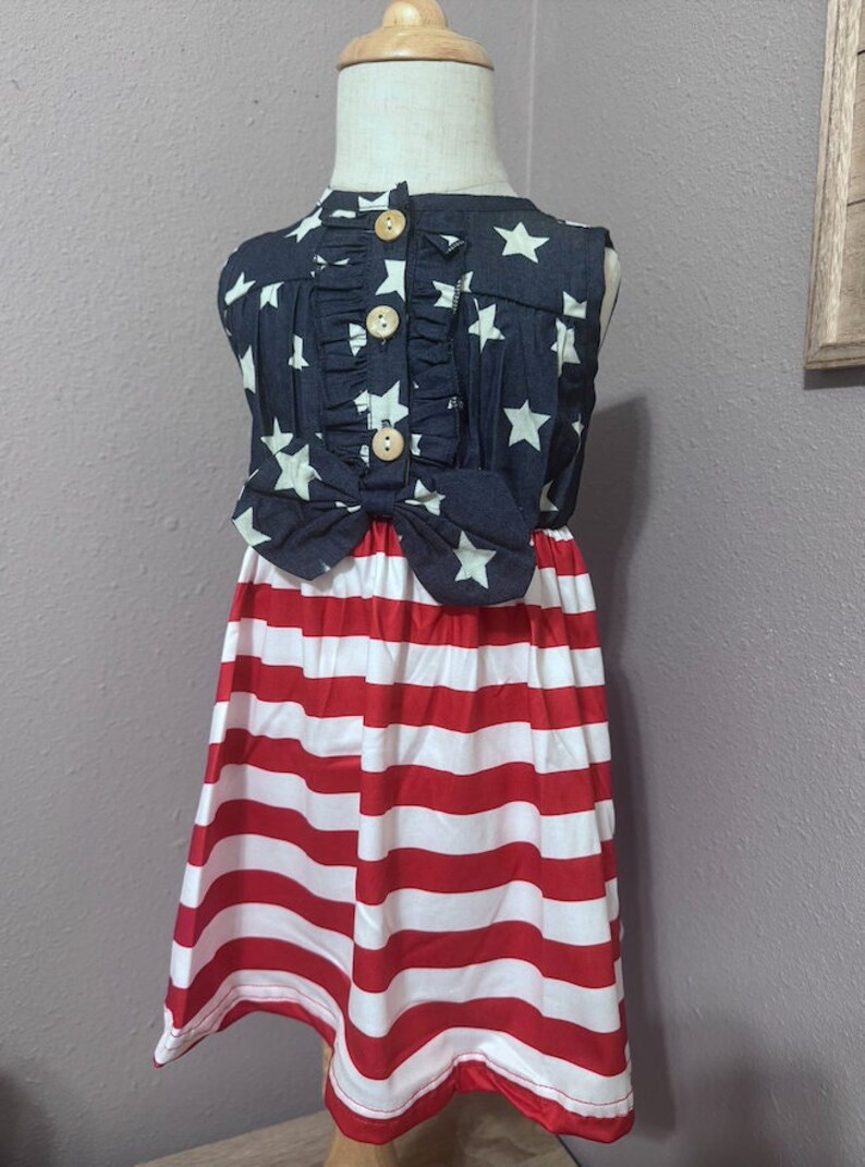 May include: A sleeveless dress for a child, featuring a blue and white star pattern on the top and red and white stripes on the bottom. The dress has a bow at the waist and wooden buttons down the front.