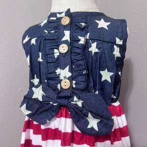 May include: A sleeveless dress with a blue and white star print top and a red and white striped bottom. The dress has a bow at the waist and wooden buttons down the back.