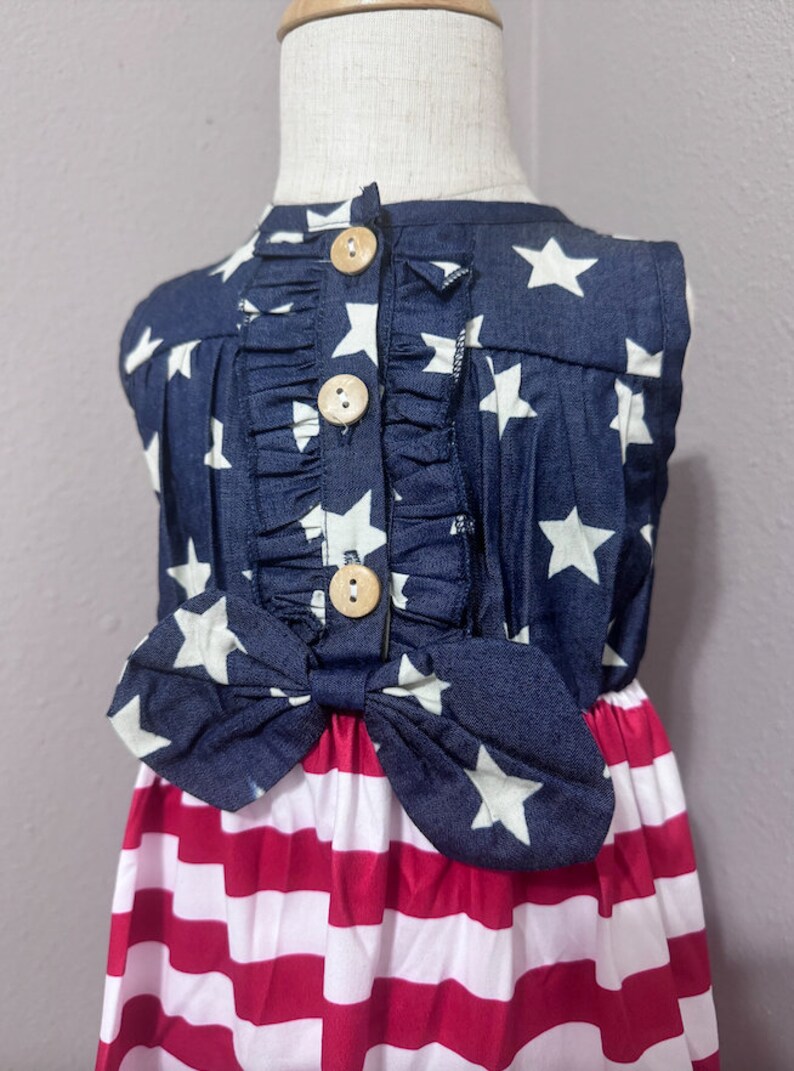 May include: A sleeveless dress with a blue and white star print top and a red and white striped bottom. The dress has a bow at the waist and wooden buttons down the back.