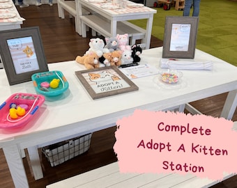 May include: A white table with a variety of items for a kitten adoption event. There are stuffed kittens, a sign that says "Adopt a Kitten!" and a sign that says "Time for a Bath". There are also two toy bathtubs with rubber ducks.