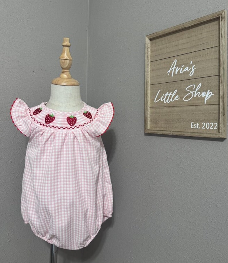 K&ouml;nnte beinhalten: Ein rosa-wei&szlig;es Gingham-Baby-Bodysuit mit gestickten Erdbeeren um den Halsausschnitt. Der Bodysuit hat kurze, gekr&auml;uselte &Auml;rmel. Der Bodysuit befindet sich auf einer Holzpuppe.