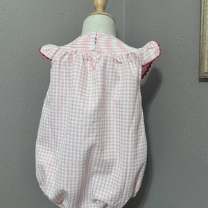 K&ouml;nnte beinhalten: Ein rosa-wei&szlig;es Gingham-Baby-Bodysuit mit roten R&auml;ndern an den &Auml;rmeln. Der Bodysuit hat einen R&uuml;schenkragen und einen Knopfverschluss auf der R&uuml;ckseite.