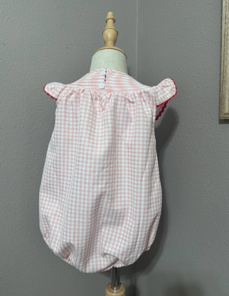 K&ouml;nnte beinhalten: Ein rosa-wei&szlig;es Gingham-Baby-Bodysuit mit roten R&auml;ndern an den &Auml;rmeln. Der Bodysuit hat einen R&uuml;schenkragen und einen Knopfverschluss auf der R&uuml;ckseite.