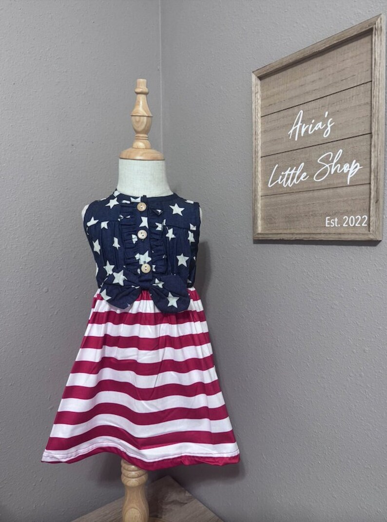 May include: A sleeveless dress with a blue and white star print top and a red and white striped skirt. The dress is on a mannequin and has a bow at the waist.