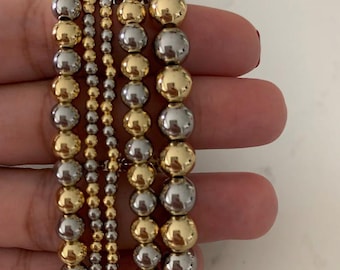 May include: Several beaded bracelets in gold and silver tones. The bracelets vary in size, with some featuring larger beads and others smaller. The beads have a polished, reflective surface, creating a shiny appearance. The bracelets are stacked together.