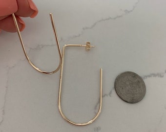 May include: A pair of gold-toned hoop earrings with a unique U-shaped design. The earrings are shown on a white marble surface next to a US quarter for size reference.
