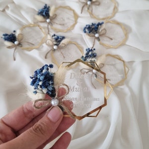 May include: Gold hexagonal acrylic wedding favour with a blue and white dried flower arrangement and the text "Dulek & Murat Nisan Hitirasi 09.04.2023"