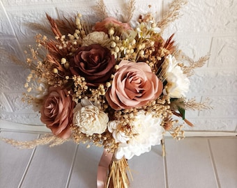 May include: A bouquet of dried flowers in shades of brown, pink, and white. The bouquet includes roses, dried grasses, and other dried flowers. The bouquet is tied with a pink ribbon.