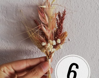 May include: A small, dried floral arrangement featuring wheat, grasses, and small white flowers. The bouquet is tied with a ribbon and held in a hand. The number 6 is in a white circle in the background.