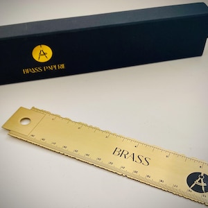 May include: A gold-colored metal ruler with a circular hole at one end, featuring the word BRASS and a compass logo printed on the surface. The ruler is accompanied by a long, dark blue rectangular box labeled with the BRASS PAPERIE logo.