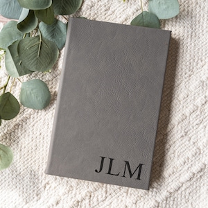 May include: A gray journal with the initials "JLM" embossed in black. The journal has a textured cover and is lying on a white, textured surface. Green leaves are in the background.