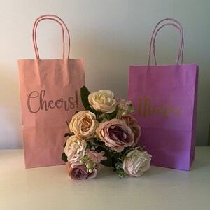 May include: Two pink and purple gift bags with gold lettering. The pink bag says "Cheers!" and the purple bag says "Maria". A bouquet of pink and purple flowers is in front of the bags.