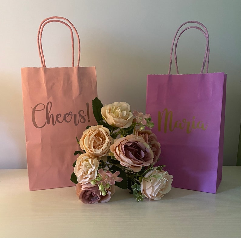 May include: Two pink and purple gift bags with gold lettering. The pink bag says "Cheers!" and the purple bag says "Maria". A bouquet of pink and purple flowers is in front of the bags.