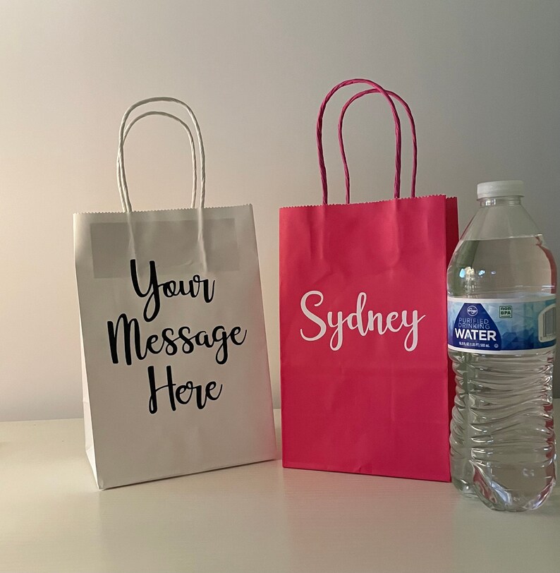 May include: Two white paper gift bags with handles. One bag has the text "Your Message Here" printed on it. The other bag is pink and has the name "Sydney" printed on it. A clear plastic bottle of water is in the foreground.