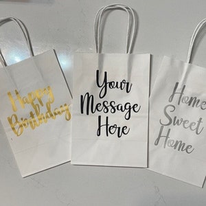 May include: Three white paper gift bags with handles. The bags have different gold and silver lettering. One bag says "Happy Birthday", one says "Your Message Here", and one says "Home Sweet Home".
