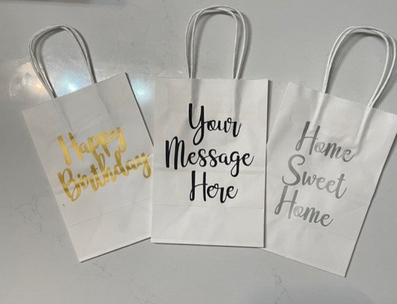 May include: Three white paper gift bags with handles. The bags have different gold and silver lettering. One bag says "Happy Birthday", one says "Your Message Here", and one says "Home Sweet Home".