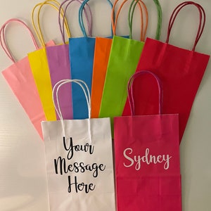 May include: Assortment of colourful paper gift bags with twisted paper handles. The bags are in various colours, including pink, yellow, lavender, light blue, orange, green, and red. Two bags have personalised text: one says "Your Message Here" and the other says "Sydney".