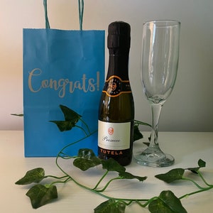 May include: A blue gift bag with the word "Congrats!" printed on it, a small bottle of Prosecco, and a champagne flute. The bottle is labelled "Prosecco" and "Tutela".