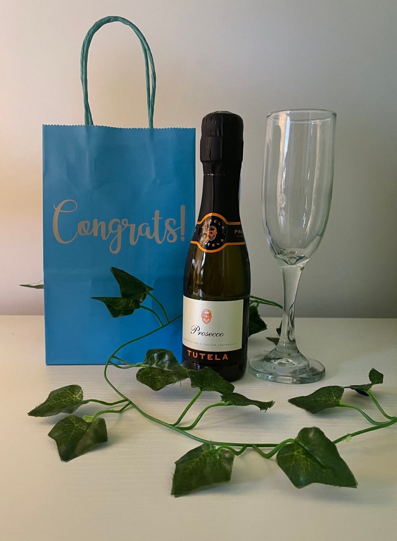 May include: A blue gift bag with the word "Congrats!" printed on it, a small bottle of Prosecco, and a champagne flute. The bottle is labelled "Prosecco" and "Tutela".