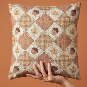 May include: A square throw pillow featuring a diamond-patterned tapestry weave in shades of brown, tan, and cream. The design alternates between light-colored diamonds containing embroidered motifs of brown acorns and yellow maple leaves.