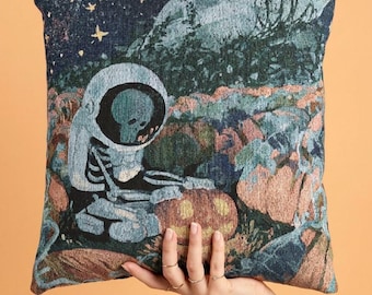 Può includere: Un cuscino quadrato con un design a arazzo intrecciato che raffigura uno scheletro con casco da astronauta, seduto in un campo di zucche sotto un cielo notturno blu scuro stellato. La scena include zucche arancioni e formazioni rocciose.