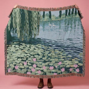 May include: A woven throw blanket featuring a landscape scene with a large weeping willow tree, a pond with two white swans, and numerous green lily pads with pink water lily flowers. The blanket has a fringed edge on all four sides.