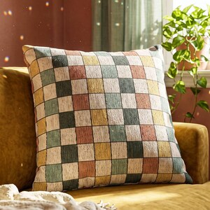May include: A square throw pillow featuring a multi-colored checkered pattern in shades of muted green, dusty rose, mustard yellow, and cream. The fabric has a visible woven texture, and the pillow is positioned on a mustard yellow sofa.