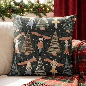 May include: A square throw pillow featuring a dark green fabric with a woven pattern of nutcracker figures, ballerinas, and stylized Christmas trees. The design includes small white stars and tan ribbon banners scattered across the dark green background.
