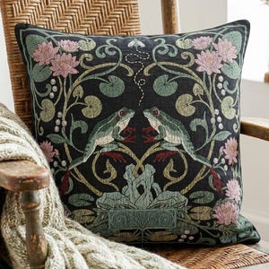May include: A square throw pillow with a black background featuring a symmetrical woven design of two green frogs facing each other, surrounded by pink water lilies, green lily pads, and swirling vines, with a small white insect above them.