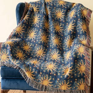 May include: A woven throw blanket featuring a repeating pattern of golden yellow suns against a deep navy blue background. The blanket has a grid-like design with small stars between the suns and finished edges with decorative multi-colored fringe.
