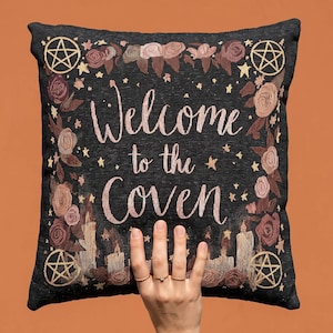 Può includere: Un cuscino quadrato nero con il testo ricamato Welcome to the Coven in corsivo rosa chiaro. Il design include motivi floreali, piccole stelle, quattro pentagrammi negli angoli e candele accese lungo il bordo inferiore del cuscino.