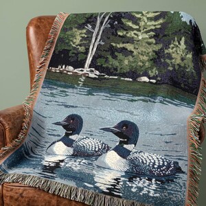 May include: A woven throw blanket featuring a detailed scene of two loons swimming on a calm lake with a dark green forest shoreline in the background. The blanket has a decorative fringe border with multi-colored threads along the edges.