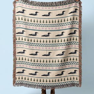 May include: A woven throw blanket featuring a repeating pattern of black and white loons, rows of green pine trees, and horizontal bands of light purple floral motifs on a cream background, finished with a decorative fringed edge on all sides.