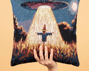 Può includere: Un cuscino quadrato con un design a arazzo intrecciato che raffigura uno spaventapasseri in un campo di notte. Un grande UFO fluttua sopra, proiettando un fascio di luce dorata sullo spaventapasseri, sotto un cielo blu scuro con una luna.