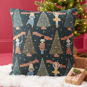 May include: A square throw pillow featuring a dark green fabric with a woven pattern of nutcracker figures, ballerinas, and stylized Christmas trees. The design includes horizontal peach-colored banners interspersed among the festive holiday motifs.