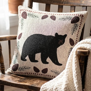 May include: A square throw pillow featuring a woven design of a solid black bear centered on a light beige background. The border is decorated with a repeating pattern of dark brown pinecones and green pine needles in a rectangular frame.