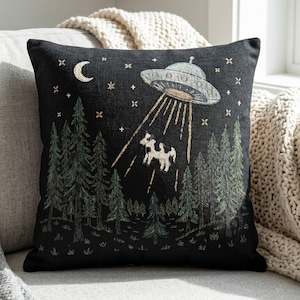 May include: A square black throw pillow featuring an embroidered scene of a silver UFO hovering over a dark forest of green pine trees. A white cow is being lifted into the air by a beam of light from the craft, with a crescent moon and stars above.