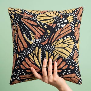 May include: A square throw pillow featuring a repeating monarch butterfly wing pattern in shades of orange, yellow, and black. The fabric has a textured weave, and the design includes white spots along the black wing borders.