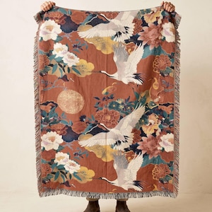 Puede incluir: Una manta tejida rectangular con un fondo de color óxido y un patrón repetitivo de grullas blancas en vuelo, grandes flores en tonos blanco, amarillo y rojo oscuro, y un elemento circular dorado que recuerda a la luna.