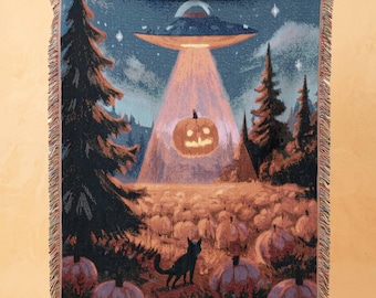 Può includere: Una coperta tessuta che raffigura un cielo notturno blu scuro con un disco volante che proietta luce su una zucca illuminata in un campo. Un gatto nero si trova in primo piano tra alberi di pino e numerose zucche con bordi sfrangiati.