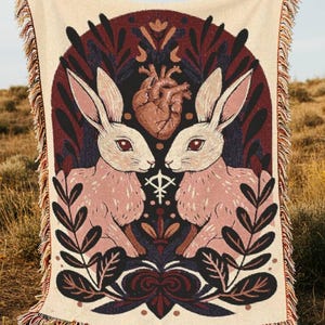 May include: Cream-coloured woven throw blanket featuring a design with two pink rabbits facing each other, a brown heart, and dark red and black floral elements. The blanket has a fringed edge and is displayed outdoors.