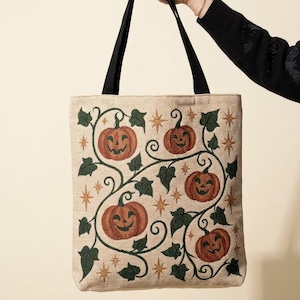 Può includere: Una borsa tote beige con tracolle nere caratterizzata da un motivo ricamato di zucche arancioni, viticci con foglie verde scuro e piccoli accenti a forma di stella dorata sulla superficie frontale della borsa in tessuto rettangolare.