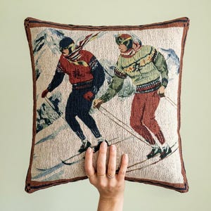 May include: A square throw pillow featuring a woven tapestry design of two skiers on a snowy mountain slope. The left skier wears a red sweater and dark pants, while the right skier wears a green patterned sweater and red pants, framed by a brown border.