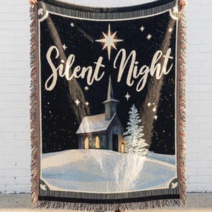 Könnte beinhalten: Eine gewebte Wohndecke mit einem dunklen Nachthimmel, einem großen Stern über einer Kirche und einer verschneiten Landschaft. Der Schriftzug Silent Night steht in weißer Schreibschrift oben. Die Decke hat an allen Seiten Fransen.