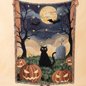 May include: A woven throw blanket featuring a nighttime Halloween scene with a large yellow full moon, flying bats, and bare black trees. A black cat sits in the foreground among orange jack-o'-lanterns and a gray tombstone marked with RIP.