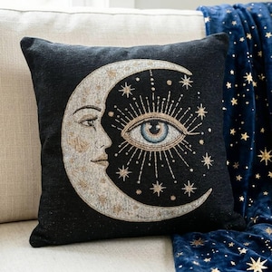 May include: A square black throw pillow featuring a woven design of a crescent moon with a human face profile. The moon is rendered in shades of silver and gold, with a central blue eye surrounded by radiating lines and small gold stars.